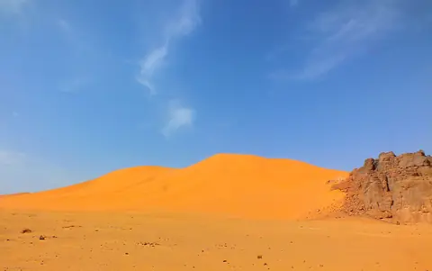 Orange sanddyn och en brun klippformation under blå himmel i Tassili n'Ajjer i Sahara