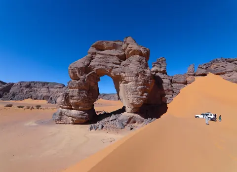 Naturlig klippbåge vid en orange sanddyn under klarblå himmel i Tadrart Acacus i Sahara, med en terrängbil vid dynens fot
