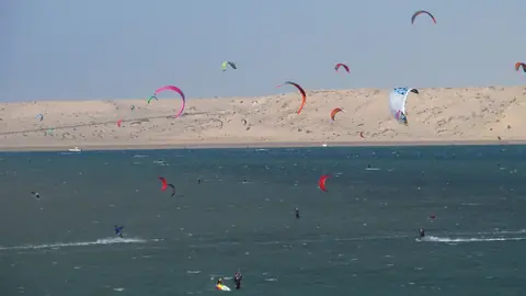 Färgglada drakar från kitesurfare över lagunen vid Dakhla, med ljusa sanddyner på andra sidan