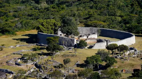 Den höga, rundade stenmuren kring Great Enclosure i ruinstaden Great Zimbabwe bland träd och gräs