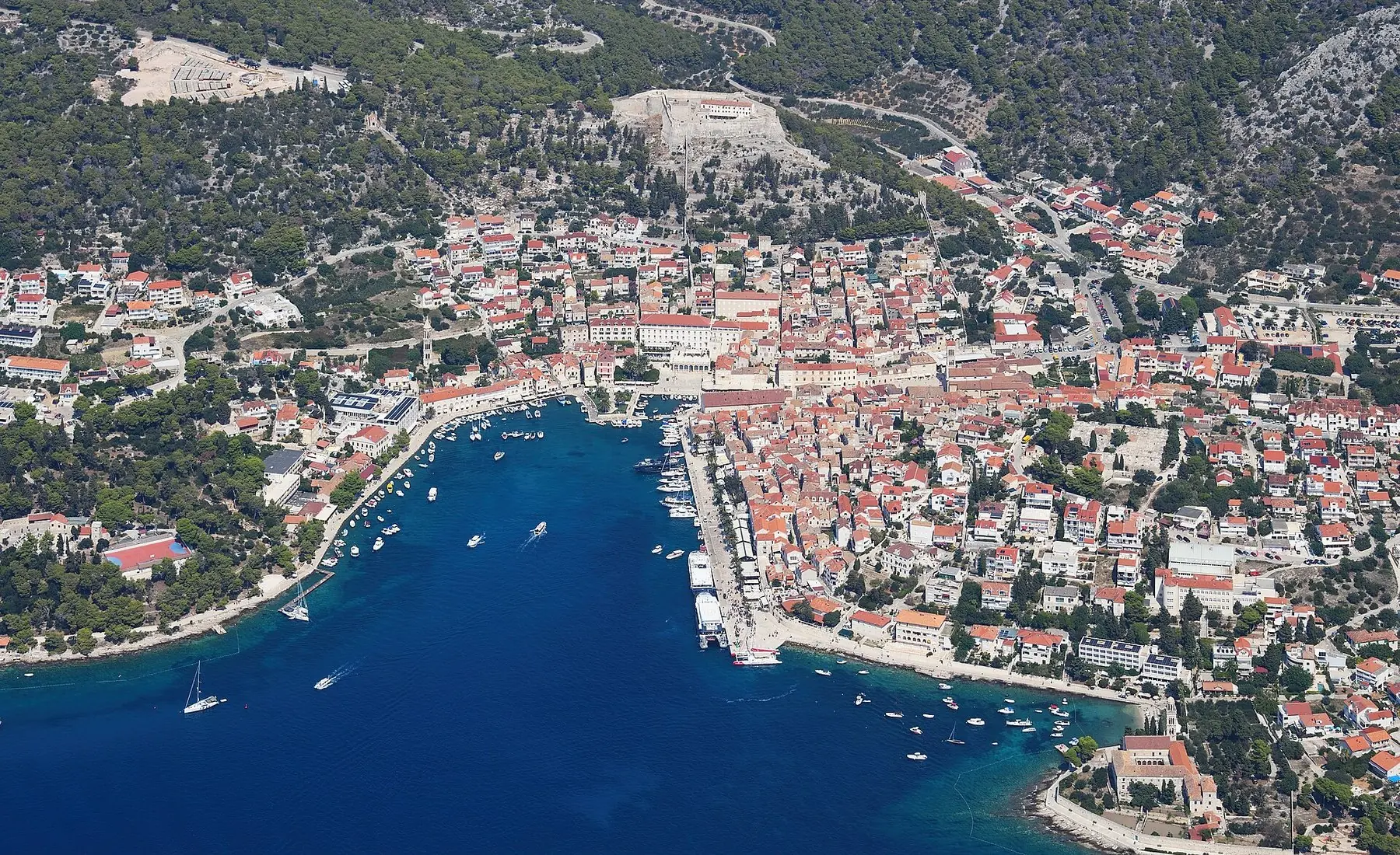 Staden Hvar med röda tak kring den djupblå hamnviken och gröna kullar, sett från luften
