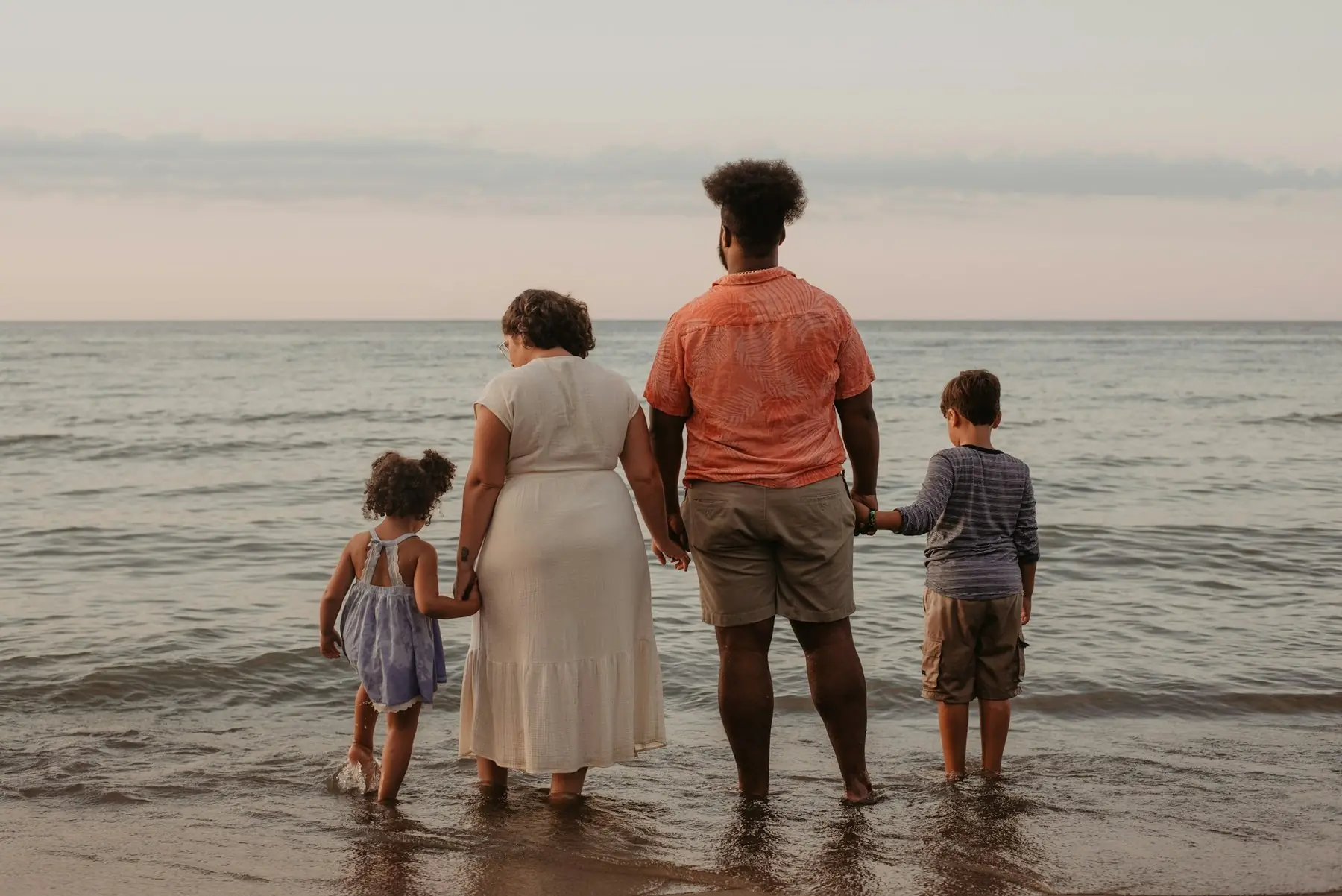 Familj med två barn står i strandkanten och ser ut över havet, sedda bakifrån