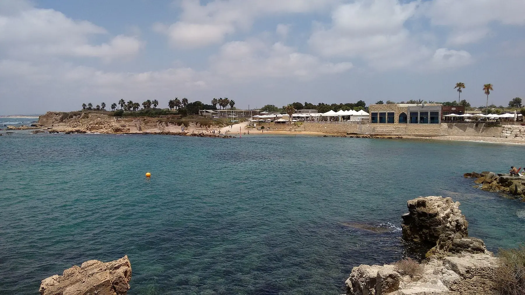 Turkost Medelhavsvik med klippor och den gamla hamnen i Caesarea under ljusa moln