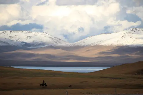 En ryttare på gyllene betesmark ovanför sjön Song-Kul med snöklädda berg under drivande moln i Kirgizistan