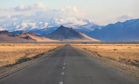 En rak väg genom gyllene stäpp mot snöklädda berg i Bayan-Ölgii i västra Mongoliet