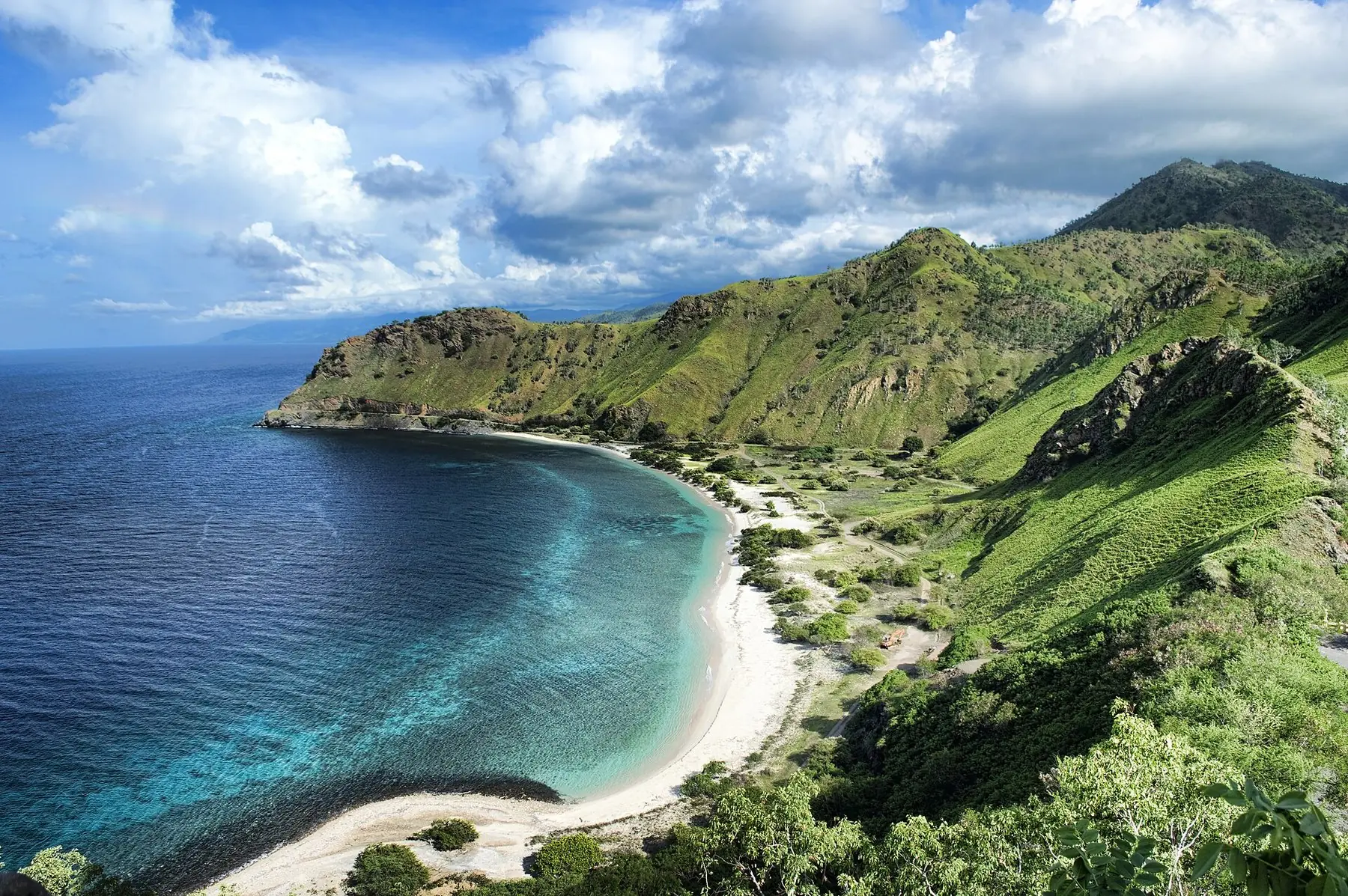 Vit sandstrand i en turkos vik under gröna kullar vid Areia Branca utanför Dili i Östtimor