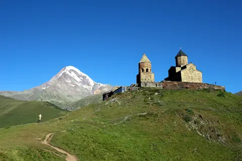 Gergetis treenighetskyrka på en grön bergsrygg med den snöklädda toppen Kazbek bakom under blå himmel