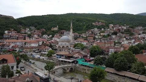 Moské med kupol och minaret vid floden och en stenbro, omgiven av röda tak och skogklädda berg i Prizren