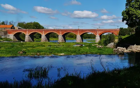 Den långa tegelbron över floden Venta i Kuldīga med gröna stränder under blå himmel