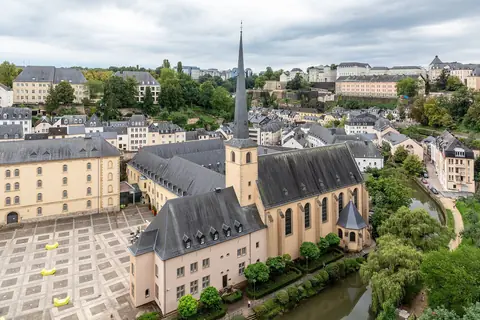 Klosterkyrkan i stadsdelen Grund med spetsigt torn, omgiven av grönska och gamla hus i Luxemburgs stad