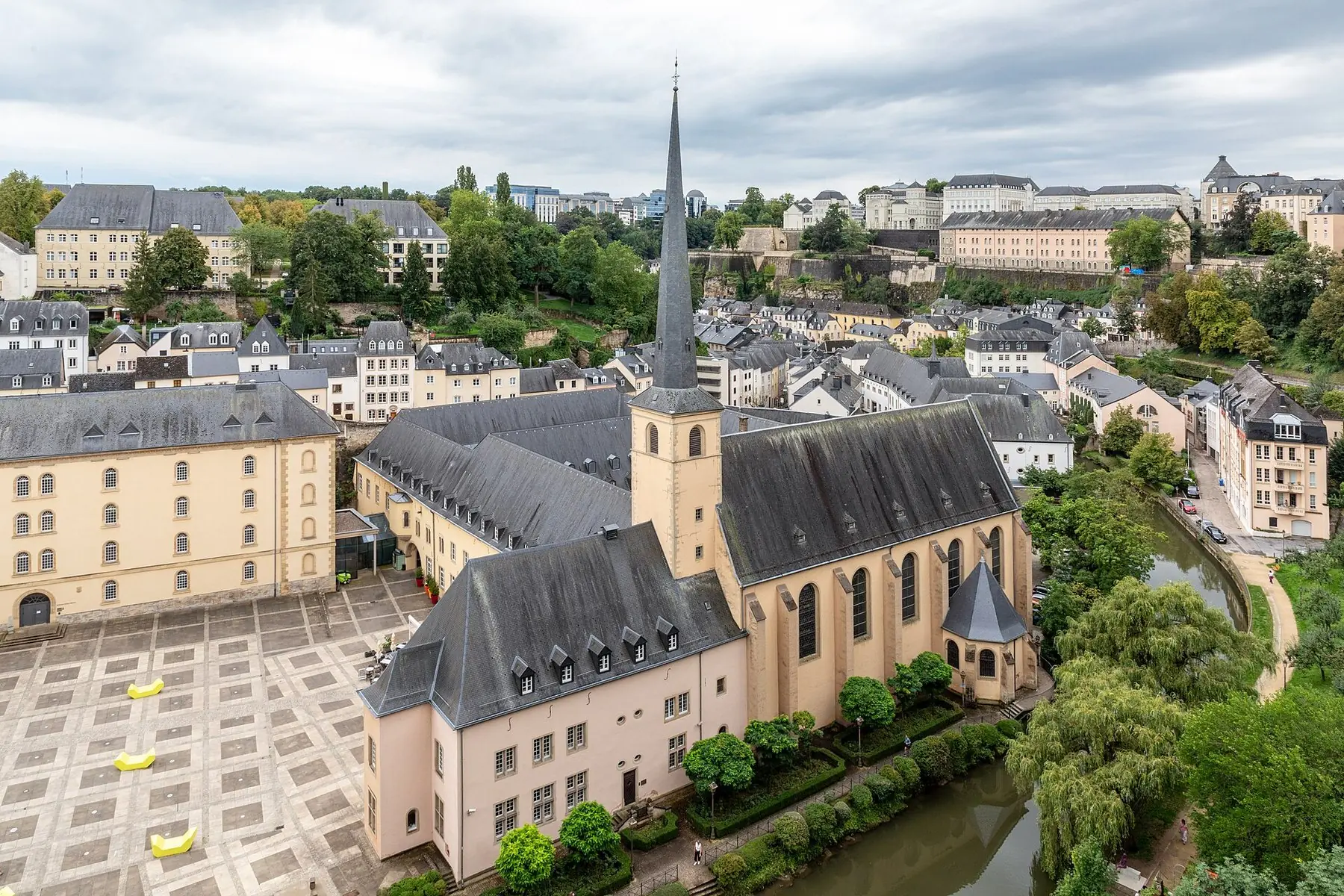 Klosterkyrkan i stadsdelen Grund med spetsigt torn, omgiven av grönska och gamla hus i Luxemburgs stad