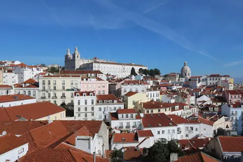 Alfamas vita hus och röda tegeltak under blå himmel i Lissabon, med kyrkan São Vicente de Fora i bakgrunden
