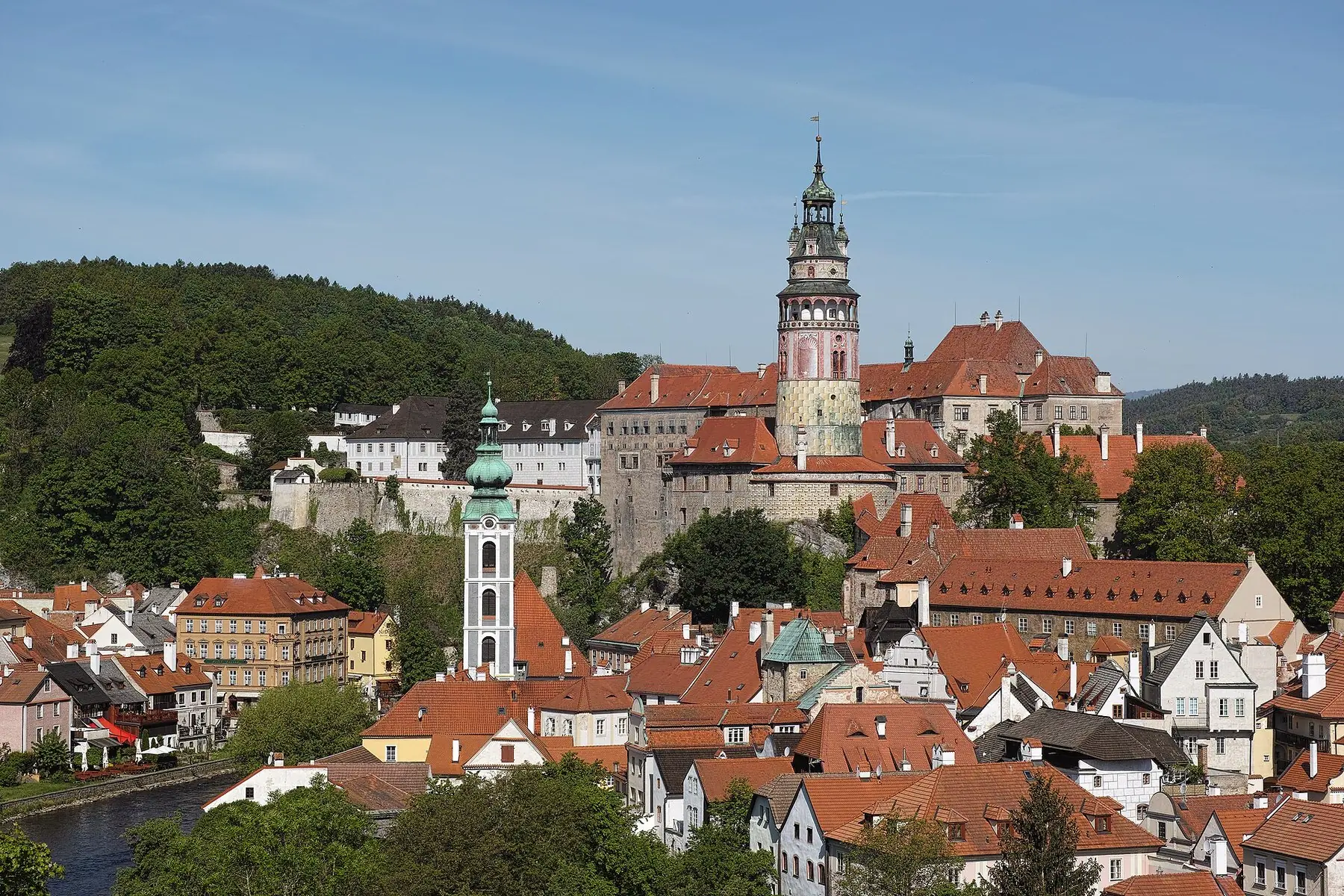 Röda tegeltak och slottstornet i Český Krumlov mellan gröna kullar under blå himmel