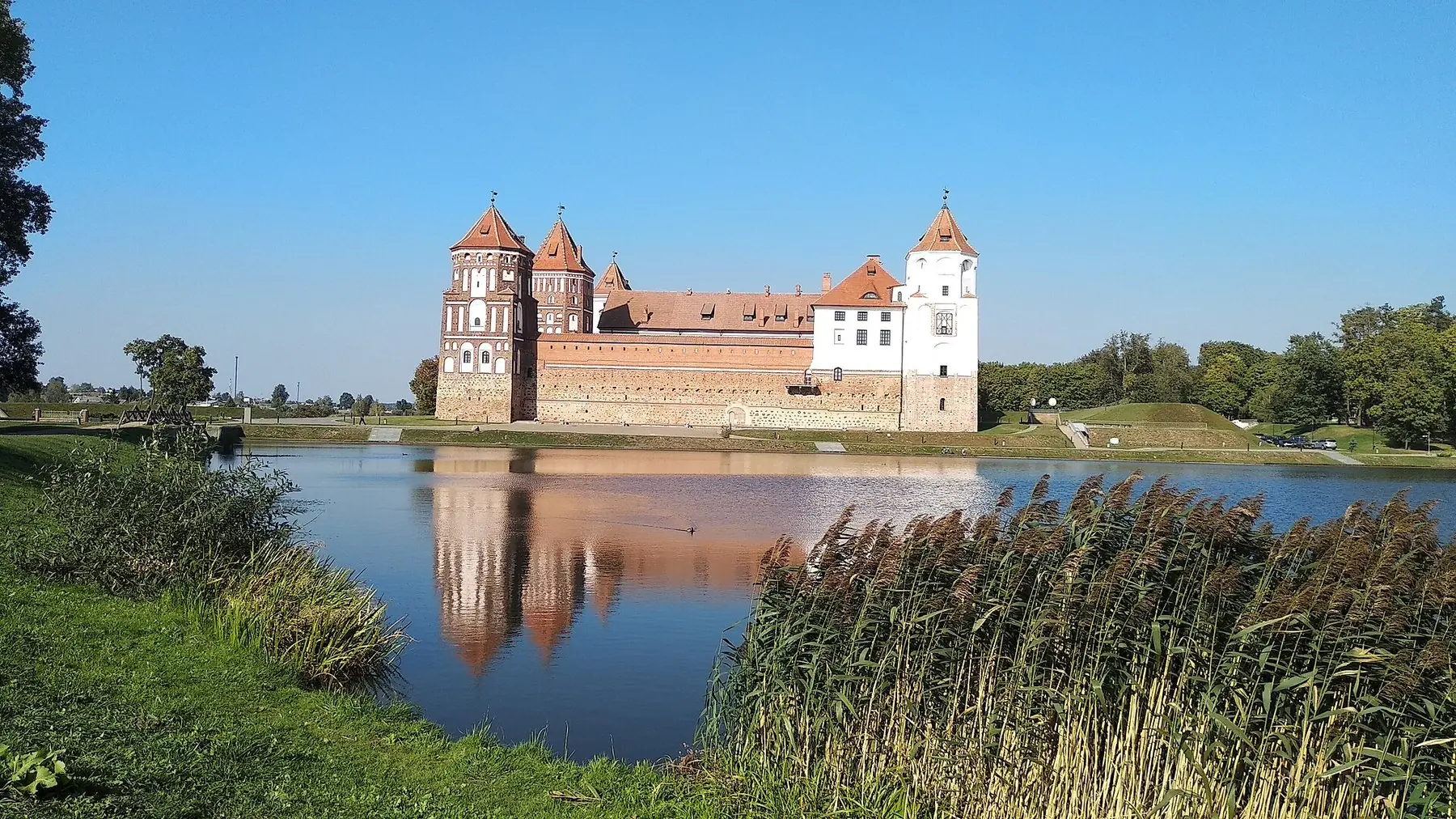Mirs slott med röda tak och torn speglat i en sjö kantad av vass i Belarus