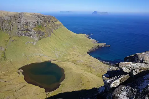 Grön klippplatå med en liten sjö ovanför ett djupblått hav vid Hvannhagi på Suðuroy