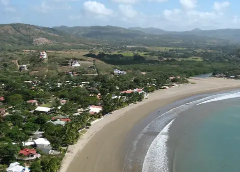 Den böjda sandstranden i San Juan del Sur med hus bland träden och gröna kullar