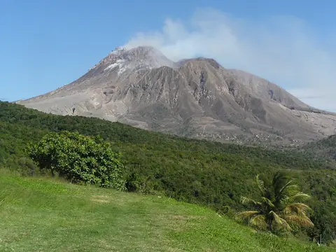 Vulkanen Soufrière Hills med grå sluttningar bakom gröna ängar och palmer