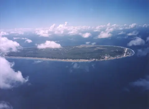 Den runda ön Nauru omgiven av ett smalt rev i det blå havet, sedd från ett flygplan ovanför molnen