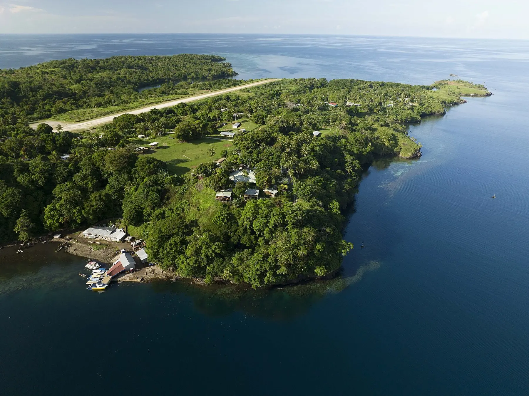 Skogklädd udde med byn Tufi och en landningsbana vid djupblått vatten i en av Tufis fjordar, sett från luften
